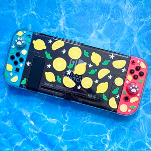 Könnte beinhalten: Eine schwarze Nintendo Switch Konsole mit einem benutzerdefinierten Design mit gelben Zitronen, grünen Blättern und weißen Blüten auf einem blauen und roten Hintergrund. Die Konsole hat einen blauen Joy-Con Controller links und einen roten Joy-Con Controller rechts. Der Text "Nintendo Switch" ist auf der Konsole sichtbar.