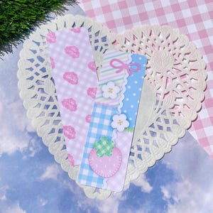 May include: Two decorative bookmarks with pastel patterns and embellishments. One bookmark features a pink gingham pattern with the text "CHU CHARMS". The other has a blue gingham pattern, a strawberry, and floral accents. They rest on a white lace doily.