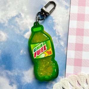 May include: A green, bottle-shaped keychain with the text "GMR FUEL" in white and red, and "Cheez dust" in yellow. The keychain has a silver clasp and is filled with glitter.