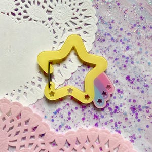 May include: A yellow star-shaped keychain with a rainbow accent and small star cutouts. The keychain has a metal clasp and is set against a background of glitter and lace doilies.