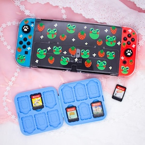 May include: A blue and black Nintendo Switch console with a clear case featuring a frog and strawberry design. The console is open and shows a blue game card case with multiple slots for game cartridges. Two game cartridges are visible, one with a red and white label and the other with a blue and white label.