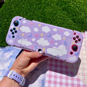 May include: A lavender handheld gaming console featuring a pattern of white clouds, yellow stars, blue crescent moons, and pink hearts. The device includes two detachable controllers with black joysticks and buttons, all encased in a protective shell.