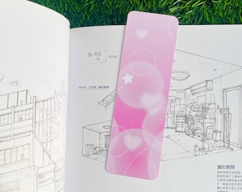 May include: A pink bookmark featuring a white star and hearts. The background is a gradient of pink and white with white circles.