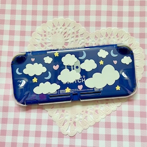 Peut inclure: Une coque bleue pour Nintendo Switch avec un dos transparent et un motif de nuages blancs, d'étoiles et de lunes. La coque porte le logo Nintendo Switch sur le devant.