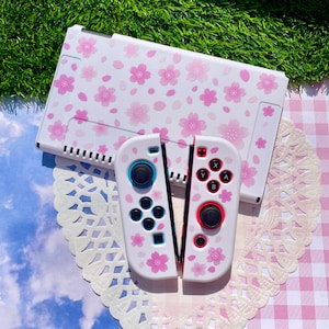 May include: A white gaming console dock and two matching controllers featuring a pink cherry blossom pattern. The left controller has blue accents around the joystick and buttons, while the right controller features red accents around the joystick and buttons.