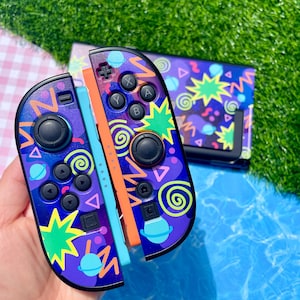 Könnte beinhalten: Ein Paar Videospiel-Controller und eine Konsolen-Dockingstation mit dunkelvioletter Basis und einem lebendigen 90er-Jahre-Muster aus neongrünen Sternen, gelben Zickzacklinien, blauen Planeten und violetten Spiralen. Die Controller haben schwarze Tasten.