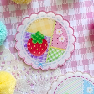 May include: A circular, pastel-colored patch with a scalloped edge. The patch features a strawberry, floral, and plaid design. The strawberry is red with green leaves. The background is a pink and white gingham pattern.