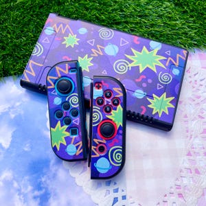 May include: A video game console and two controllers featuring a purple background with a repeating pattern of light blue planets, yellow zig-zag lines, green starburst shapes, and white spirals. The controllers have black joysticks and buttons.