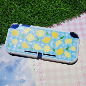 May include: A handheld gaming console protective case featuring a light blue and white gingham checkered pattern decorated with yellow lemon slices, whole lemons, and small green leaf accents, finished with a white border and dark blue accents.