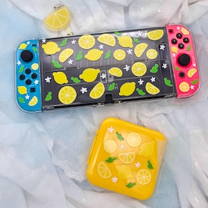 May include: A blue and pink Nintendo Switch console with a lemon-themed design. The console has a clear case with yellow lemon stickers and a pink Joy-Con controller. The console is on a white surface.