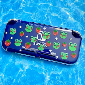 May include: A blue Nintendo Switch game console with a clear protective case. The case is decorated with a pattern of green frogs and red strawberries. The text "Nintendo Switch" is printed on the case.