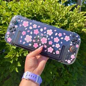 May include: A clear protective case for a Nintendo Switch console, decorated with pink cherry blossom designs. The case has a dark background and features the Nintendo Switch logo. The case is held against a backdrop of green foliage.