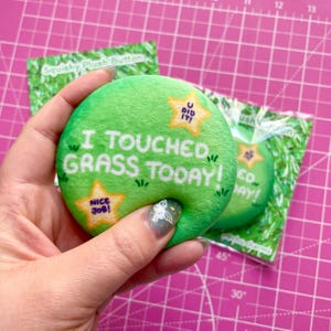 Pode incluir: Botão de pelúcia verde com o texto "I Touched Grass Today!" e uma estrela com o texto "U Did It!" e outra estrela com o texto "Nice Job!"