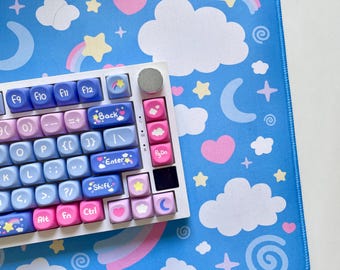 Peut inclure: Un clavier blanc avec des touches de couleur pastel représentant un arc-en-ciel, des étoiles et des nuages. Le clavier est posé sur un tapis de souris bleu à motifs de nuages et d'étoiles.