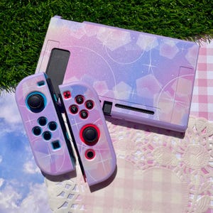 May include: A gaming console dock and two controllers featuring a pastel purple and blue gradient finish with a glittery texture. The surface is decorated with white starbursts and geometric pentagon shapes, creating a celestial aesthetic.