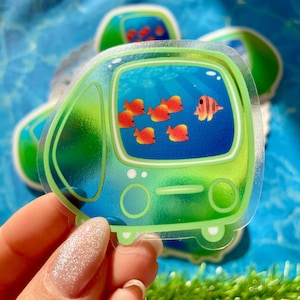 May include: A clear sticker featuring a cartoon aquarium scene. The sticker has a green border and depicts orange fish swimming in blue water. One fish has pink stripes. The sticker is held in a hand against a blue background.