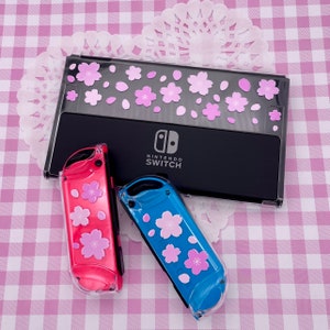 Puede incluir: Dos estuches de juego Nintendo Switch de plástico transparente con diseños florales rosas y azules. Los estuches están decorados con flores y pétalos de cerezo rosa. El texto "NINTENDO SWITCH" está impreso en la caja negra.