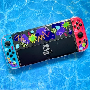 May include: A clear plastic case for a Nintendo Switch console with a colorful 90s-inspired design featuring geometric shapes, swirls, and stars. The case is pink and blue with black accents. The Nintendo Switch logo is visible on the front of the case.