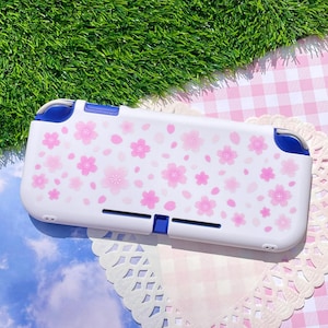 May include: A white handheld gaming console case featuring a repeating pattern of light and dark pink cherry blossom flowers. The case has blue accents on the top shoulder buttons and a central blue rectangular port cover on the bottom edge.