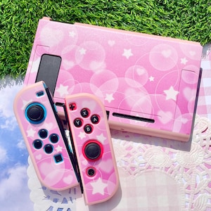 May include: A pink gaming console dock and two matching controllers featuring a pattern of white stars, hearts, and translucent circles. The controllers include black analogue sticks, directional buttons, and the standard A, B, X, and Y action buttons.
