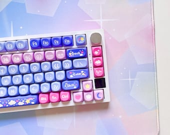 May include: A white keyboard with pastel-colored keycaps featuring various symbols and designs, including stars, hearts, and clouds. The keyboard is on a pastel blue and pink mousepad with a geometric pattern.
