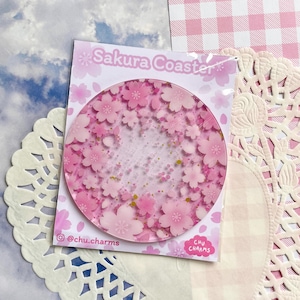 Puede incluir: Un posavasos Sakura redondo y transparente con flores de cerezo rosas y detalles de purpurina dorada. El posavasos está sobre una tarjeta blanca con el texto "Sakura Coaster" y el identificador de Instagram "@chu.charms".