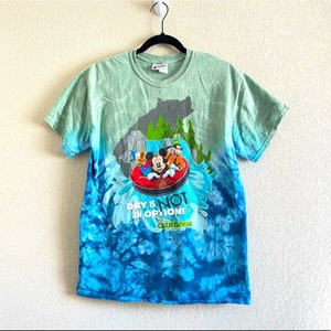Puede incluir: Camiseta teñida con un diseño de Mickey Mouse, el Pato Donald y Goofy en una atracción acuática. La camiseta tiene un degradado azul y verde con el texto "DRY IS NOT AN OPTION!" y "California Adventure".