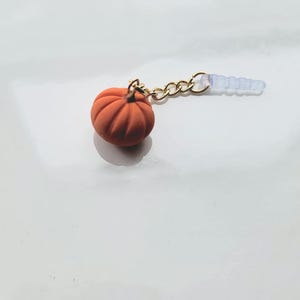 May include: A small, orange pumpkin charm with a gold-colored chain and a clear, plastic earbud dust plug. The pumpkin has detailed ridges and is set against a white background. The charm is likely for a phone or other device.