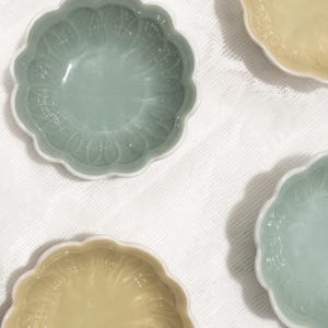 May include: Four small, decorative ceramic bowls with a scalloped edge and a floral pattern. Two bowls are a pale green, and two are a pale yellow. The bowls are on a white, textured surface.