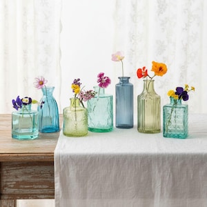 Op de afbeelding: Een verzameling van zes kleine glazen vazen in diverse tinten blauw en groen, elk met unieke texturen en vormen. Deze transparante vazen bevatten kleine, kleurrijke wilde bloemen, waaronder roze, oranje, gele en paarse bloesems.