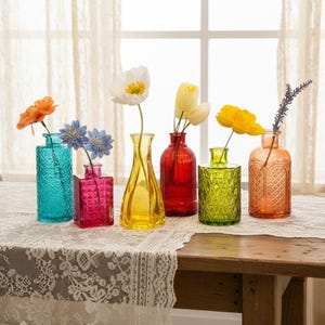 Könnte beinhalten: Eine Sammlung von sechs kleinen Glasvasen in verschiedenen Farben, darunter Türkis, Magenta, Gelb, Rot, Limettengrün und Bernstein. Jede Vase hat eine einzigartige Form und Textur und enthält jeweils einen Stiel mit künstlichen Blumen.