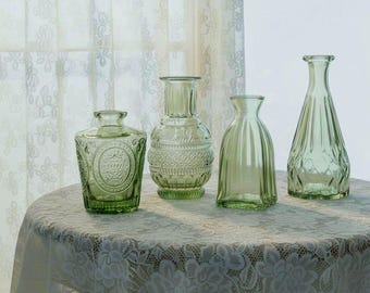 May include: A collection of four light green glass vases with distinct shapes and textures. The set includes a short round vase with an embossed circular pattern, a medium vase with a diamond grid, a fluted vase, and a tall tapered vase.