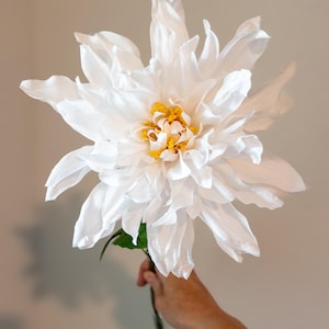 Peut inclure: Une grande fleur artificielle blanche avec de multiples couches de pétales délicats. Le centre de la fleur présente un groupe d'étamines jaunes. La tige est verte et une seule feuille verte est visible. La fleur est tenue sur un fond neutre.