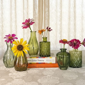 May include: A collection of six small green glass vases in various shapes and textures, each holding a single flower. The vases are arranged on two stacked books titled Flower Color Theory and Cultivated, featuring pink, yellow, and orange blooms.