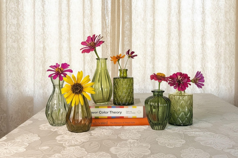 May include: A collection of six small green glass vases in various shapes and textures, each holding a single flower. The vases are arranged on two stacked books titled Flower Color Theory and Cultivated, featuring pink, yellow, and orange blooms.
