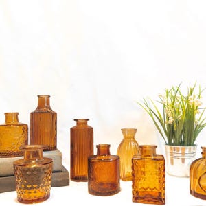 Op de afbeelding: Een assortiment amberkleurige glazen flessen in verschillende vormen en texturen, waaronder vierkante, rechthoekige en afgeronde ontwerpen. Twee lange, gedraaide kaarsen en een kleine potplant zijn ook aanwezig.