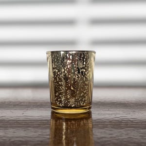 Może przedstawiać: Mały, złoty świecznik z nakrapianym wzorem. Szkło ma metaliczne wykończenie, odbijające światło. Świecznik stoi na ciemnej drewnianej powierzchni. Ma około 7 cm wysokości.