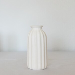 Pu&ograve; includere: Un vaso di ceramica bianca con un design a coste. Il vaso ha un collo stretto e una base arrotondata. Le costole verticali corrono lungo la lunghezza del vaso, creando una superficie strutturata. Il vaso &egrave; su una superficie bianca.