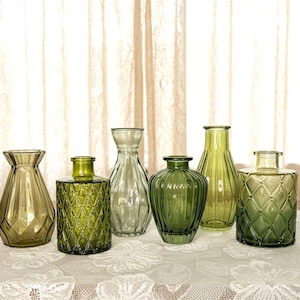 May include: A collection of six glass vases in various shades of green. The set includes vessels with diverse textures, such as diamond-patterned surfaces, vertical ribbing, and smooth finishes, all featuring narrow necks and wide bases.