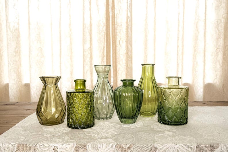 May include: A collection of six glass vases in various shades of green. The set includes vessels with diverse textures, such as diamond-patterned surfaces, vertical ribbing, and smooth finishes, all featuring narrow necks and wide bases.