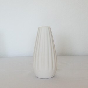 Pu&ograve; includere: Un vaso in ceramica bianca, a coste, a forma di goccia. Il vaso presenta scanalature verticali che vanno dalla base all'apertura. Lo sfondo &egrave; una superficie bianca.