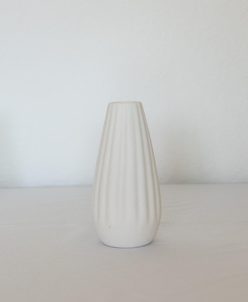 Pu&ograve; includere: Un vaso in ceramica bianca, a coste, a forma di goccia. Il vaso presenta scanalature verticali che vanno dalla base all'apertura. Lo sfondo &egrave; una superficie bianca.