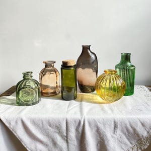 May include: A collection of six glass bottles and vases in various shapes and colors, including a small green embossed bottle, a peach-toned square bottle, a dark green bottle with a cork, a tall brown bottle, a round yellow ribbed vase, and a tall green ribbed vase.
