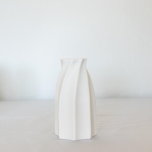 Pu&ograve; includere: Un vaso in ceramica bianca con un design unico e attorcigliato. Il vaso ha una base ampia e si assottiglia verso l'alto, con una superficie strutturata e scanalata. Il colore neutro e la forma moderna lo rendono un oggetto decorativo versatile.