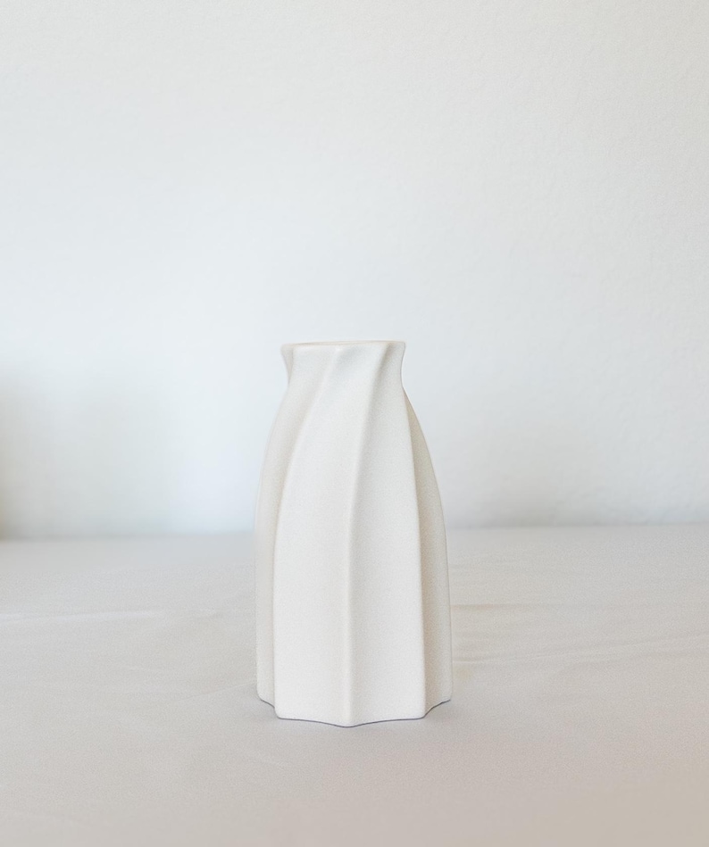 Pu&ograve; includere: Un vaso in ceramica bianca con un design unico e attorcigliato. Il vaso ha una base ampia e si assottiglia verso l'alto, con una superficie strutturata e scanalata. Il colore neutro e la forma moderna lo rendono un oggetto decorativo versatile.