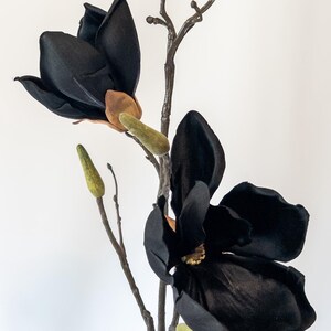 May include: A black artificial magnolia flower with a brown stem and green buds. The large, velvety petals of the flower are fully open, showcasing the intricate details. The flower is a striking decorative element.