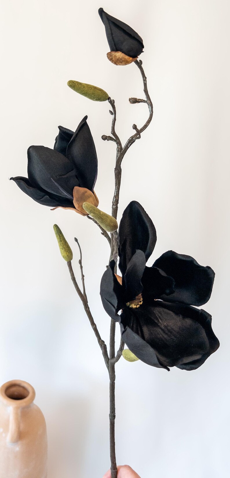 May include: A black artificial magnolia flower with a brown stem and green buds. The large, velvety petals of the flower are fully open, showcasing the intricate details. The flower is a striking decorative element.
