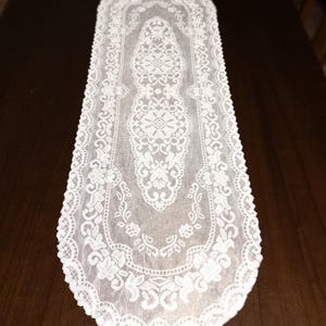 May include: A white lace table runner with an intricate floral and geometric pattern. The runner is rectangular with a scalloped edge, and is placed on a dark brown wooden surface. The lace design is detailed and delicate, adding a touch of elegance.