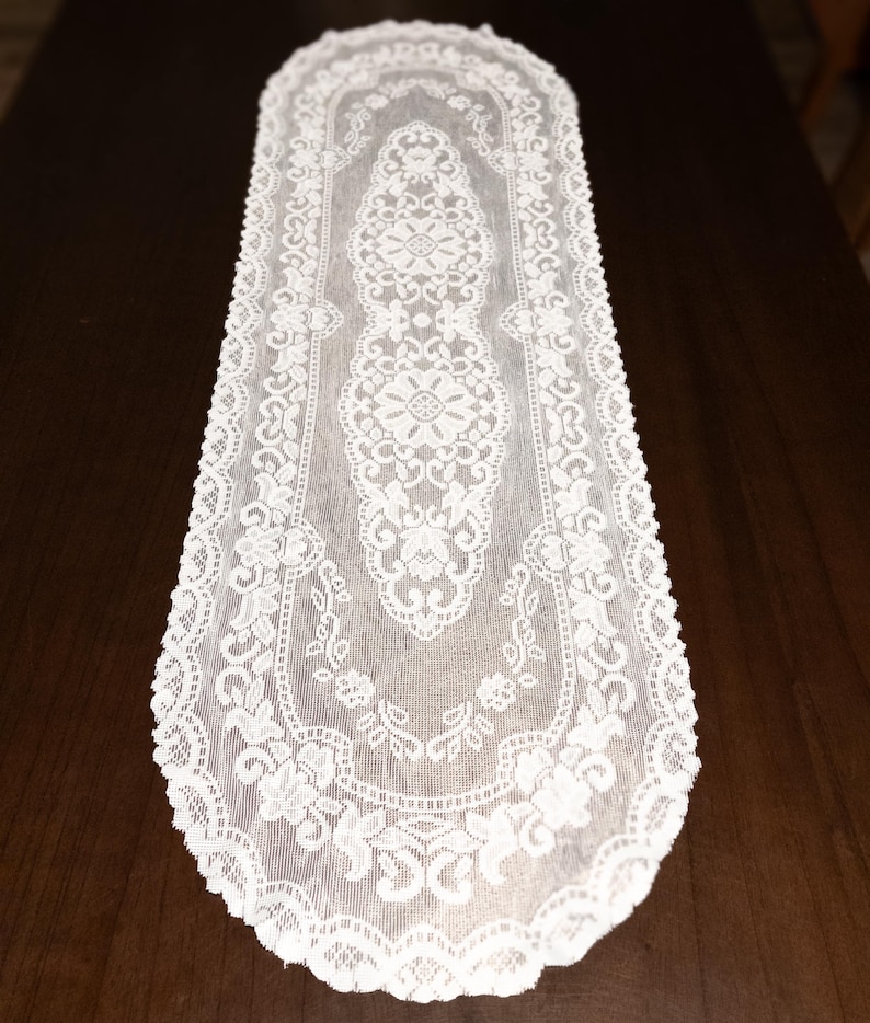 May include: A white lace table runner with an intricate floral and geometric pattern. The runner is rectangular with a scalloped edge, and is placed on a dark brown wooden surface. The lace design is detailed and delicate, adding a touch of elegance.
