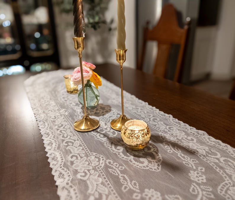 May include: A dining table is set with a white lace runner, gold candlesticks, and a small vase of flowers. Two tall gold candlesticks hold brown and green candles. A small glass votive and a gold-rimmed candle holder are also on the table.